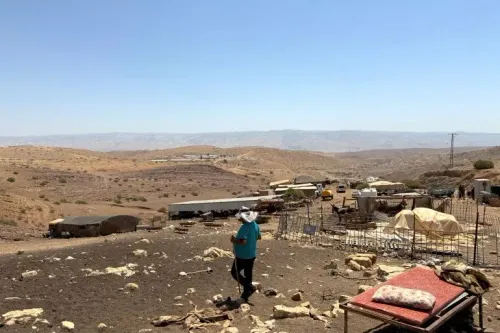 مشهد يظهر شخصًا يسير في منطقة قاحلة بالضفة الغربية، مع وجود مستوطنات ومزارع في الخلفية، يعكس تأثير الاحتلال الإسرائيلي.