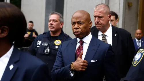 إريك آدامز، عمدة مدينة نيويورك، يظهر في صورة خارج مبنى حكومي، مع تعبير جاد، محاطًا بأفراد من الشرطة.