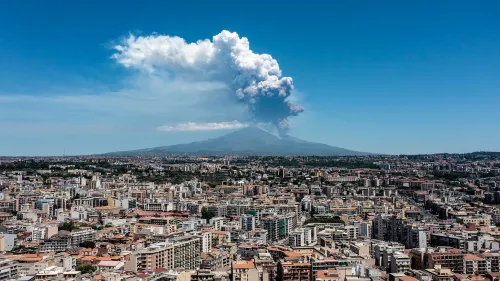 ثوران بركاني هائل في جبل إتنا، يظهر عمود من الغازات والرماد يتصاعد فوق المدينة، مع تأثيرات على الرحلات الجوية في المنطقة.