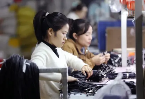 عاملتان في مصنع تقومان بتجميع كابلات الشحن، مما يعكس تأثير التعريفات الجمركية على صناعة الإلكترونيات.