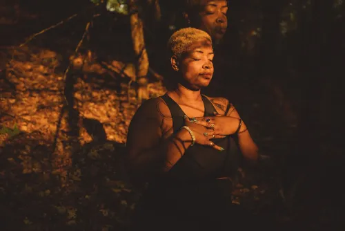 امرأة ذات بشرة داكنة تجلس في غابة مضاءة بأشعة الشمس، تتأمل وتضع يديها على صدرها. تعكس الصورة لحظة من الهدوء والتواصل مع الطبيعة.