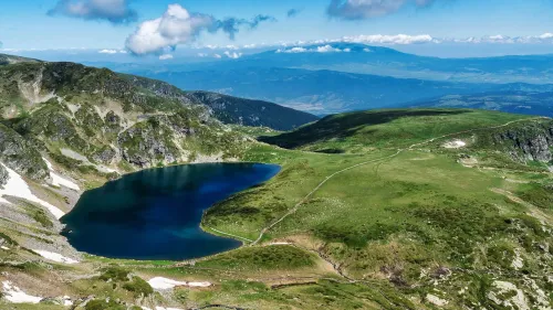 منظر بانورامي لبحيرة جليدية محاطة بالجبال الخضراء في منطقة ريلا ببلغاريا، مع سماء زرقاء وغيوم بيضاء.