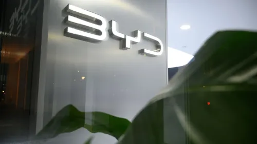 شعار شركة BYD مضيء في واجهة المبنى، مع ظهور بعض أوراق النباتات في المقدمة، مما يعكس الابتكار في صناعة السيارات الكهربائية.