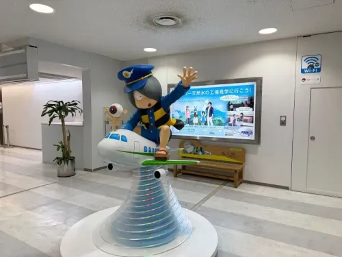 تمثال ملون لشخصية كرتونية ترتدي زي طيار، يقف على طائرة صغيرة في مطار أويتا، مع لافتات ترويجية تبرز معالم المنطقة.