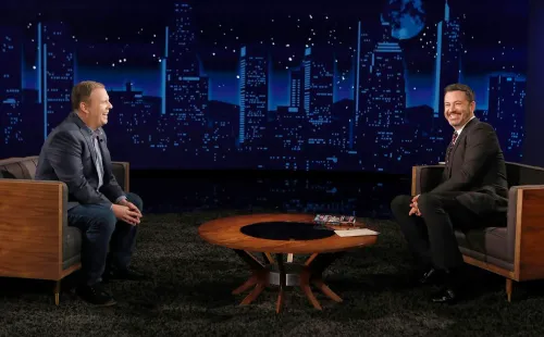 إياكونو وجيمي كيميل يجلسان في استوديو برنامج "جيمي كيميل لايف!"، مع خلفية لمدينة مضاءة ليلاً، يتبادلان الحديث والدعم في ظل التحديات الحالية.