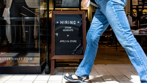 لافتة "نحن نوظف" خارج متجر، مع شخص يرتدي جينز يمشي بجانبها، تعكس زيادة فرص العمل في قطاع التجزئة.