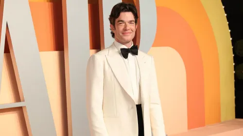 جون مولاني يرتدي بدلة بيضاء مع ربطة عنق سوداء، مبتسمًا أمام خلفية ملونة، في حدث يبرز حضوره كأب وكوميدي.