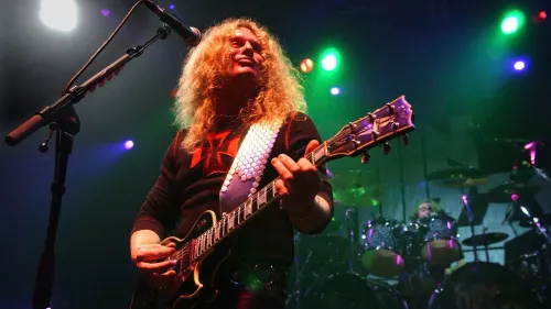 عازف الجيتار جون سايكس يؤدي على المسرح، محاطًا بأضواء ملونة، مع التركيز على موهبته الموسيقية وتأثيره في عالم الروك.