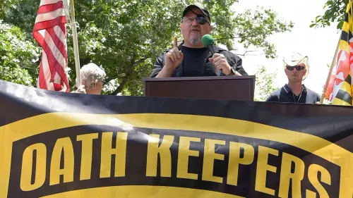 شخص يتحدث في تجمع لعدة أشخاص يحملون علم الولايات المتحدة ويظهر خلفهم لافتة تحمل اسم "حراس القسم".