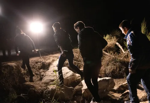 مجموعة من الشباب الغواتيماليين يسيرون في الظلام نحو الحدود، مع وجود أضواء ساطعة في الخلفية، تعكس محاولاتهم للهجرة.