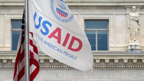 علم الوكالة الأمريكية للتنمية الدولية (USAID) يرفرف بجانب العلم الأمريكي، مع واجهة مبنى حكومي خلفهما، مما يبرز جهود الوكالة في الإغاثة والتنمية.
