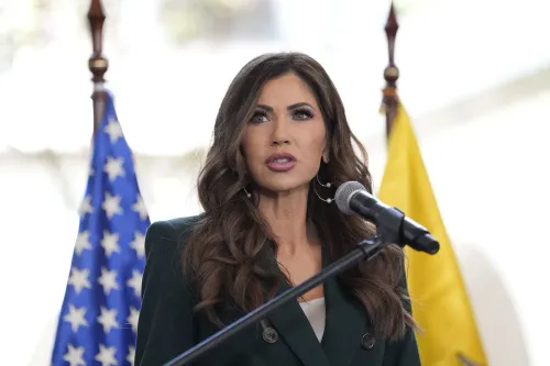 وزيرة الأمن الداخلي كريستي نويم تتحدث أمام الميكروفون، مع وجود أعلام خلفها، حول تمديد وضع الحماية المؤقتة للمهاجرين.