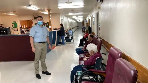 موظف في دار رعاية مسنين يقف في ممر، مع وجود مقيمين يجلسون في كراسي متحركة، مما يعكس التحديات في توفير الرعاية الصحية.