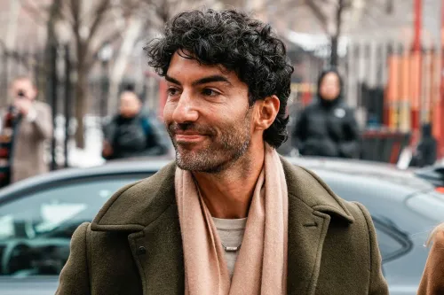 رجل ذو شعر مجعد يرتدي سترة خضراء وشالًا، يبدو مبتسمًا أثناء تواجده في منطقة خارجية مزدحمة، في سياق قضية قانونية تتعلق بالتحرش.