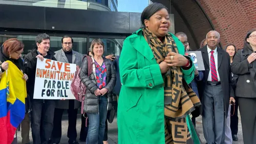 مظاهرة لدعم المهاجرين، حيث تتحدث امرأة ترتدي سترة خضراء، بينما يحمل المشاركون لافتات تطالب بالحماية الإنسانية للجميع.