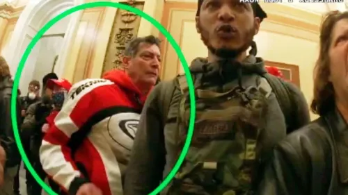 صورة تظهر مجموعة من مثيري الشغب داخل مبنى الكابيتول، مع التركيز على رجل يرتدي سترة حمراء يتفاعل مع آخرين أثناء الهجوم.