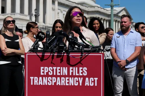 امرأة تتحدث أمام ميكروفونات في تجمع لدعم قانون الشفافية في ملفات إبشتاين، مع مجموعة من المتظاهرين خلفها.