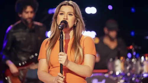 كيلي كلاركسون تغني في الحلقة رقم 1000 من برنامجها الحواري، مع فرقة موسيقية خلفها، تحت أضواء ملونة تعكس أجواء الاحتفال.