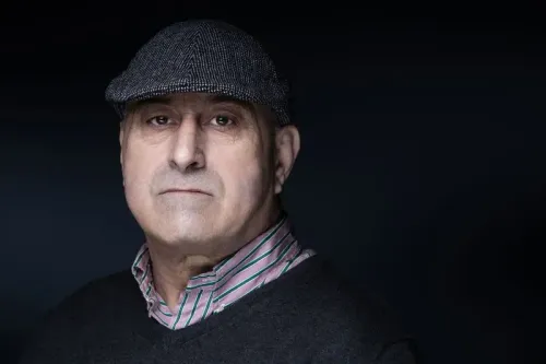 صورة لرجل مسن يرتدي قبعة رمادية وقميص مخطط، يظهر تعبيره الجاد في إضاءة خافتة، مما يعكس أجواء الجريمة في باريس.