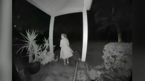 امرأة ترتدي معطفًا، تقف في مدخل منزلها ليلاً، بينما تتواجد بعض النباتات حولها، مشهد يعكس أجواء فقدان المنازل بسبب الحرائق.