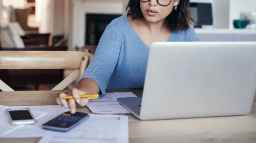 امرأة ترتدي نظارات وتستخدم حاسوبًا محمولًا، محاطة بمستندات ضريبية وآلة حاسبة، تعمل على إعداد إقرارها الضريبي قبل الموعد النهائي.