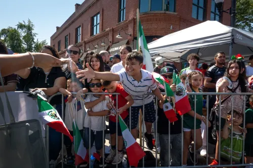 أطفال وعائلات يحملون أعلام المكسيك في احتفال يوم الاستقلال، مع ابتسامات وحماس في أجواء احتفالية في الشارع.