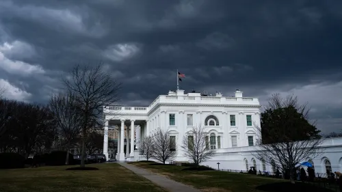 مبنى البيت الأبيض تحت سماء غائمة، مع العلم الأمريكي يرفرف فوقه. الصورة تعكس الأجواء المشحونة حول قضايا الهجرة.