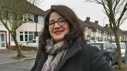 امرأة مبتسمة ترتدي نظارات وشالًا، تقف في شارع سكني محاط بالمنازل والسيارات، تعكس تأثير الذكاء الاصطناعي على المترجمين.