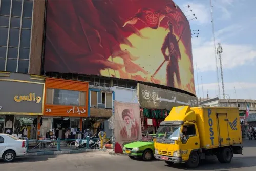 شاحنة بريد إيرانية صفراء تمر أمام مبانٍ تجارية في طهران تحت لوحة جدارية كبيرة تعكس الأوضاع الاقتصادية الصعبة في إيران.