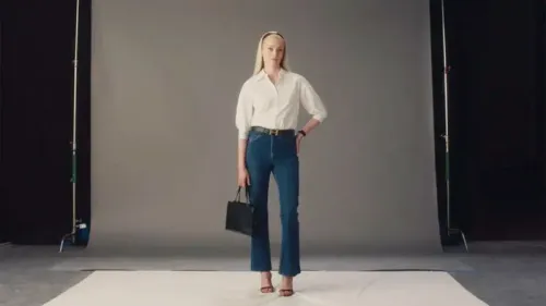 امرأة ترتدي قميصًا أبيض وسروال جينز ضيق، تحمل حقيبة سوداء، وتقف في استوديو تصوير مع خلفية رمادية، تعكس أسلوب الموضة العصري.