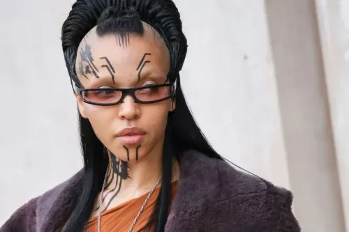 إطلالة فريدة لفنانة FKA Twigs بشعر منتفخ وحواجب مبيضة، تتزين بوشوم مؤقتة، ونظارات ملونة، تعكس أسلوبها الغريب والمستوحى من القوطية في أسبوع الموضة بباريس.