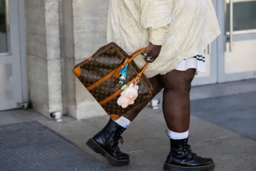 شخص يحمل حقيبة لويس فويتون مزينة بقطع ملونة، يرتدي قميصًا مخططًا وسروالًا قصيرًا، بينما يمشي أمام مكتبة نيويورك العامة.