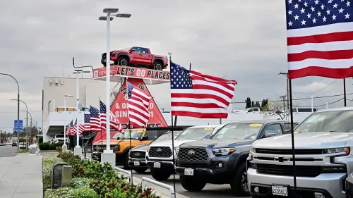 معرض سيارات أمريكي يعرض سيارات جديدة مع لافتة ترويجية، محاط بأعلام أمريكية، في ظل توقعات بزيادة الأسعار بسبب التعريفات الجمركية.