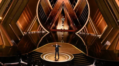 مقدم حفل الأوسكار يقف على المسرح أمام تمثال الأوسكار، مع تصميم داخلي أنيق يبرز الأضواء والديكورات الذهبية.