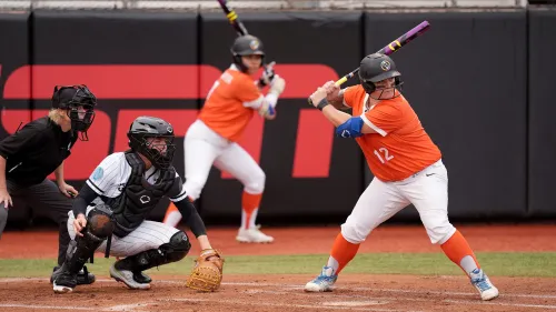 لاعبة سوفتبول ترتدي قميصًا برتقاليًا وتستعد لضرب الكرة، بينما يقف الحارس خلفها، مما يعكس أجواء المنافسة في دوري البيسبول اللينة.