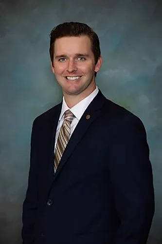 صورة لرجل مبتسم يرتدي بدلة رسمية، يظهر في خلفية رمادية، في سياق حادث اعتداء على عضو مجلس مدينة دانفيل.