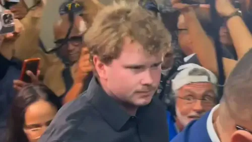 شخص شاب ذو شعر أشقر ومجعد، يرتدي قميصًا أسود، يظهر وسط حشد من الناس مع هواتف محمولة، بعد جلسة استماع في المحكمة حول قضية طالبة مفقودة.