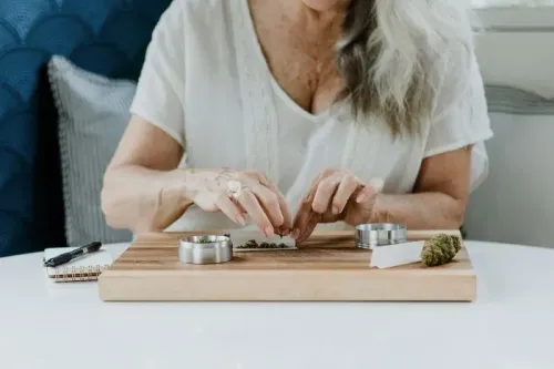 امرأة مسنّة تقوم بلف الحشيش على طاولة خشبية، مع وجود أدوات التقطيع وكمية من الأعشاب بجانبها.