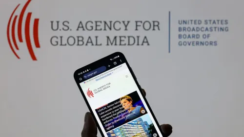 شخص يحمل هاتفًا ذكيًا يعرض موقع وكالة الولايات المتحدة للإعلام العالمي، مع شعار الوكالة في الخلفية، في سياق مناقشة حرية الصحافة.