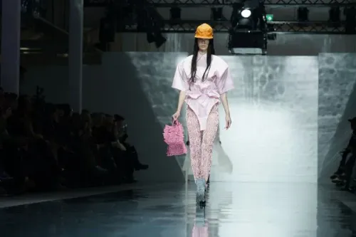 عارضة أزياء تسير على المدرج مرتدية ملابس مبتكرة من اللاتكس، تشمل قميصًا ورديًا وسروالًا شفافًا، مع قبعة برتقالية وحقيبة ملونة.