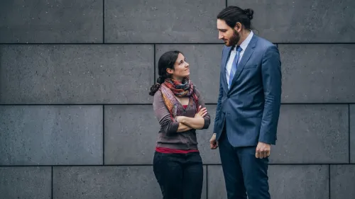 امرأة ورجل يتحدثان معًا في مكان خارجي، حيث يظهر الرجل بزي رسمي أكبر طولًا من المرأة التي ترتدي ملابس غير رسمية.