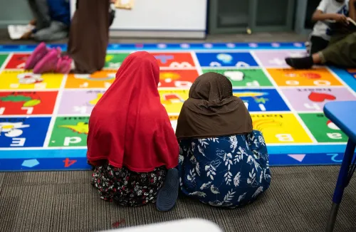 طفلتان ترتديان الحجاب تجلسان معًا على سجادة ملونة في مركز رعاية أطفال، مع وجود أطفال آخرين في الخلفية.