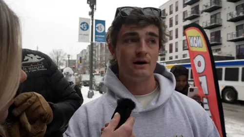 شخص يتحدث في الشارع خلال طقس ثلجي، مع لافتة تشير إلى حدث محلي، بينما تظهر مبانٍ خلفه.