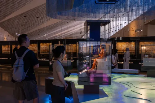 زوار يتأملون تمثالًا قديمًا داخل المتحف المصري الكبير، مع عرض للقطع الأثرية خلف زجاج شفاف في أجواء معمارية حديثة.