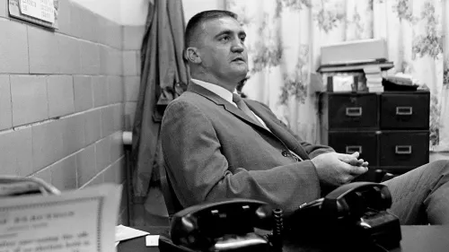 صورة تاريخية لبوفورد بوسر، المأمور الشهير، يجلس في مكتبه محاطًا بأجهزة الهاتف، مع خلفية جدارية بسيطة.