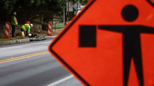 عمال بناء يرتدون سترات عاكسة يعملون في موقع لأشغال الطرق، مع وجود علامة تحذير مرورية تشير إلى العمل.