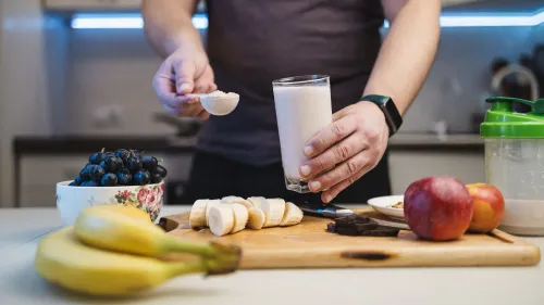 شخص يحمل ملعقة من مسحوق البروتين بالقرب من كوب من مشروب البروتين، مع فواكه مثل الموز والتفاح، مما يعكس استخدام المكملات الغذائية لبناء العضلات.