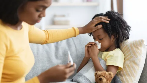 أم تعتني بابنتها المريضة على الأريكة، حيث تقيس درجة حرارتها بينما تحمل الطفلة مناديل وتبدو متعبة.