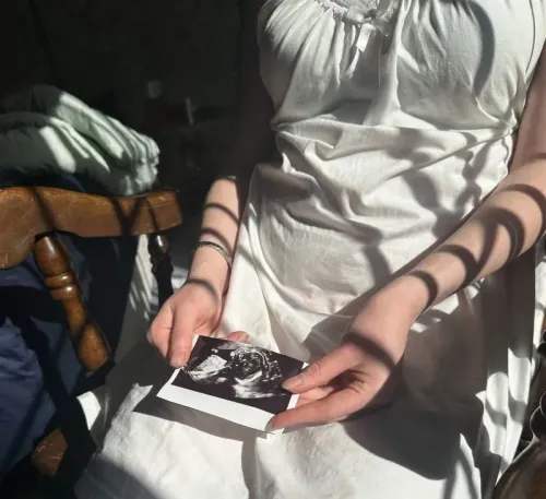 امرأة ترتدي فستانًا أبيض وتحمل صورة أشعة فوق صوتية لطفلها، بينما تسقط ظلال على ذراعيها، تعكس مشاعر القلق حول مستقبل طفلهما في ظل التوترات السياسية.