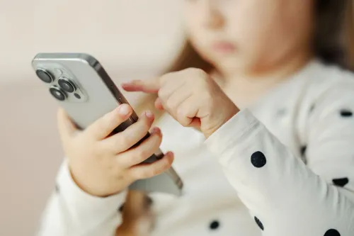 طفلة صغيرة ترتدي قميصًا مزخرفًا، تستخدم هاتفًا ذكيًا، مما يعكس قلق التقرير حول تأثير الشاشات على صحة الأطفال.